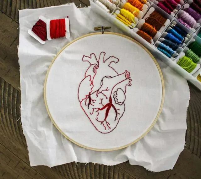 Un corazón bordado en tela blanca, listo para aprender a bordar. Hilos rojos y una caja de hilos de colores completan la escena.