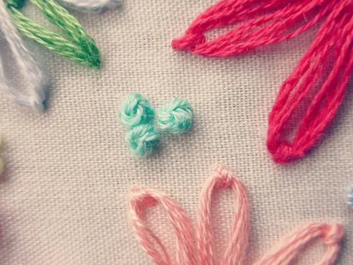 Detalle de bordado con flores de hilo. Se aprecian pétalos en rosa, rojo, azul y blanco, y un centro de nudo francés bordado en turquesa.