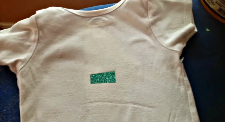 Camiseta blanca con un pequeño rectángulo verde brillante en el centro. Aprende como quitar pegatinas termoadhesivas de la ropa.