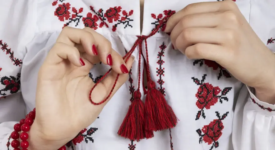 Manos atando cordones rojos en una blusa blanca con **bordado digital** de flores rojas y negras.