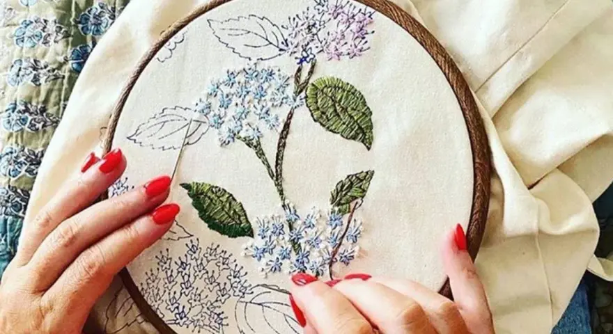 Manos con uñas rojas bordando una flor de hortensia en un bastidor. Se ve lo que se necesita para bordar: tela, hilo y aguja.