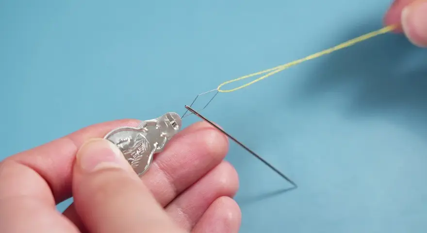 Manos usando un enhebrador de agujas para pasar hilo amarillo por el ojo de una aguja.
