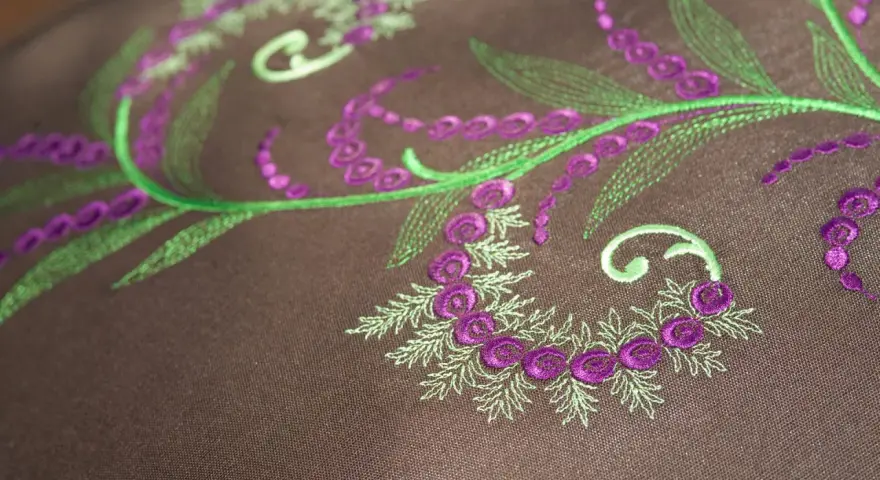 Detalles de bordado floral en tonos verdes y morados sobre tela marrón. Ideal para descargar letras para bordar a maquina y crear diseños únicos.