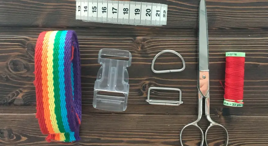 Herramientas para hacer un collar para perro: cinta métrica, cinta de arcoíris, hebilla, anillas, tijeras y hilo rojo.