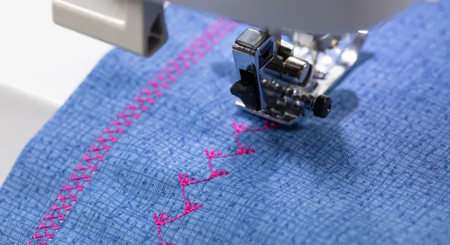 Máquina de coser bordando un patrón decorativo en **tejidos de algodón** azul con hilo rosa.