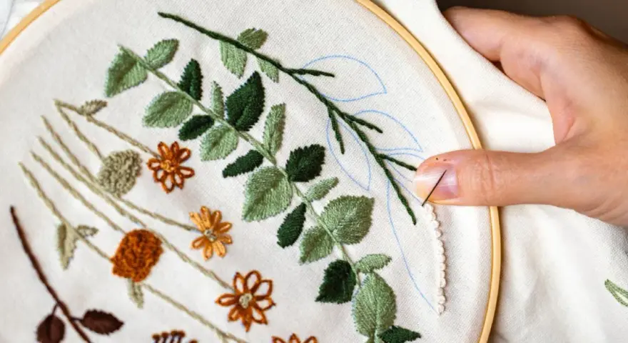 Manos creando bellas ideas para bordar: hojas verdes y flores naranjas en tela, con aguja e hilo listos para continuar.