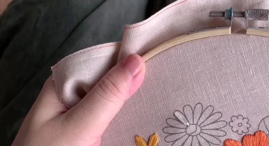 Mano sujetando un bastidor de bordar con lino para bordar. Se aprecian flores a medio bordar en tonos grises, naranjas y amarillos.