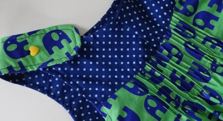 Detalle de un vestido infantil con estampado de elefantes azules sobre fondo verde. El cuello tiene un detalle de lunares azules y un botón amarillo en forma de corazón. Un adorable **patrón ranita bebé**.