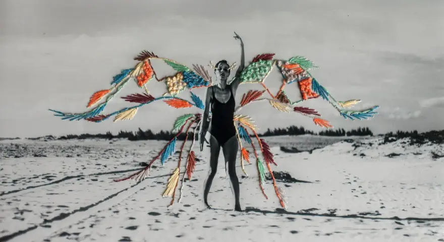 Mujer en traje de baño con alas de bordado de fotos coloridas sobre un paisaje de playa desértica.