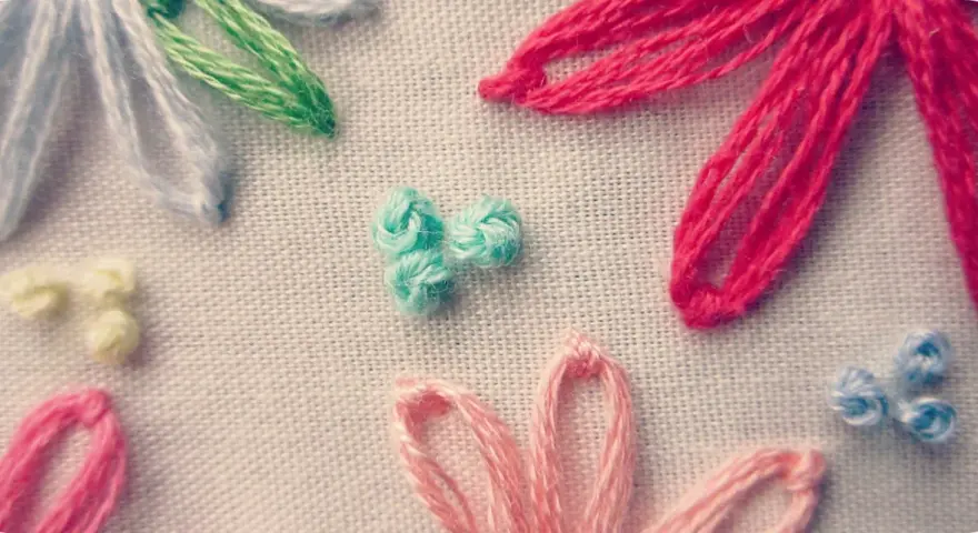Detalle de bordado con flores de hilo. Se aprecian pétalos en rosa, rojo, azul y blanco, y un centro de nudo francés bordado en turquesa.