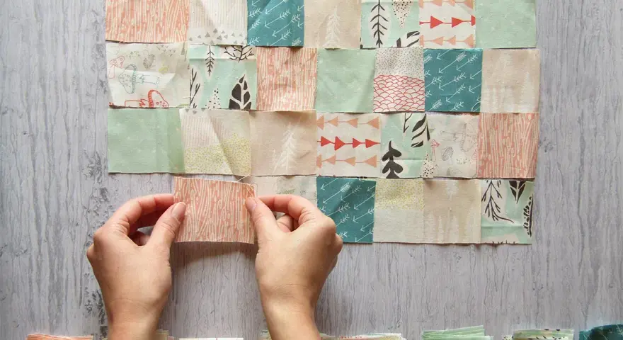 Manos uniendo retazos de tela con diseños de árboles y flechas, creando un patrón de patchwork.