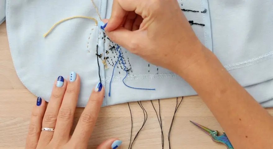 Manos con uñas decoradas bordando con hilo de coser sobre tela azul claro. Tijeras de colores y hilos negros a la vista.