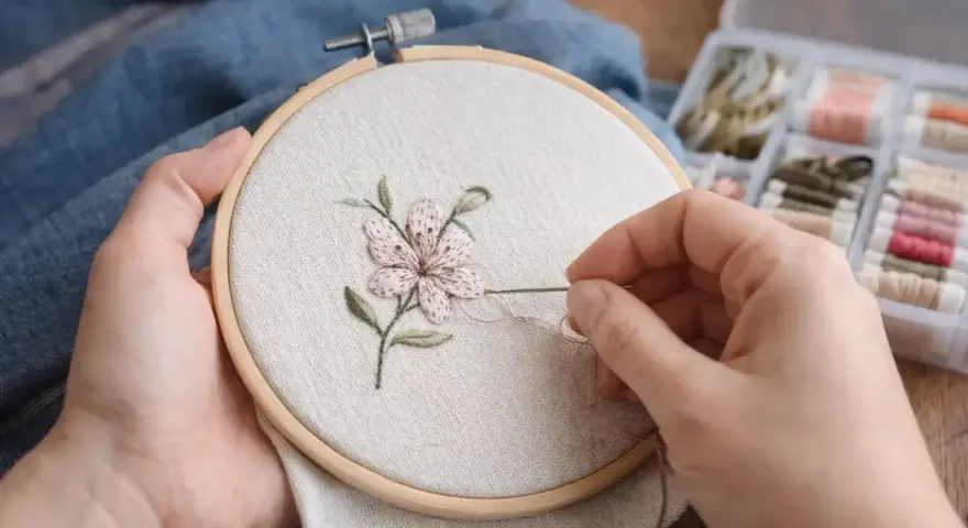 Manos delicadas bordando una flor con hilo rosa y verde sobre tela clara, usando un bastidor. A un lado, una caja con carretes de hilo.