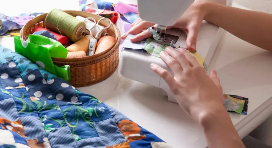 Manos cosiendo en máquina, con un costurero lleno de hilos y tijeras, y un quilt colorido.