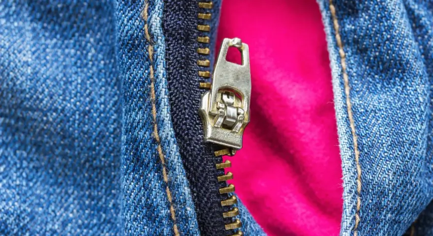 Detalle de cremallera metálica en jeans azules, con un forro rosa vibrante que se asoma. La cremallera salida de un lado revela el contraste de texturas.