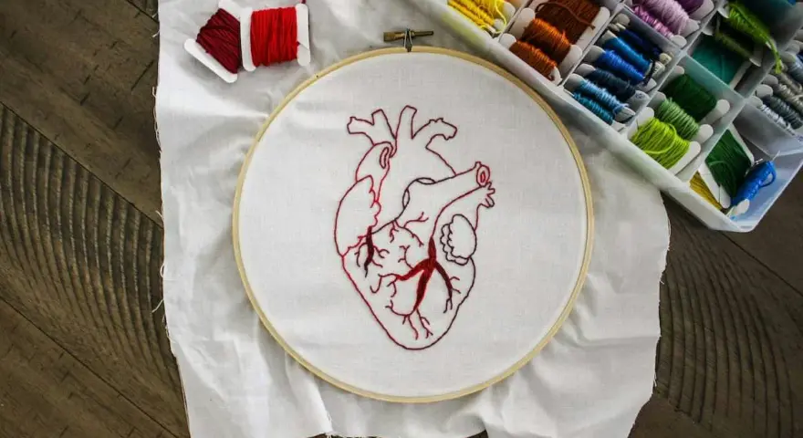 Un corazón bordado en tela blanca, listo para aprender a bordar. Hilos rojos y una caja de hilos de colores completan la escena.