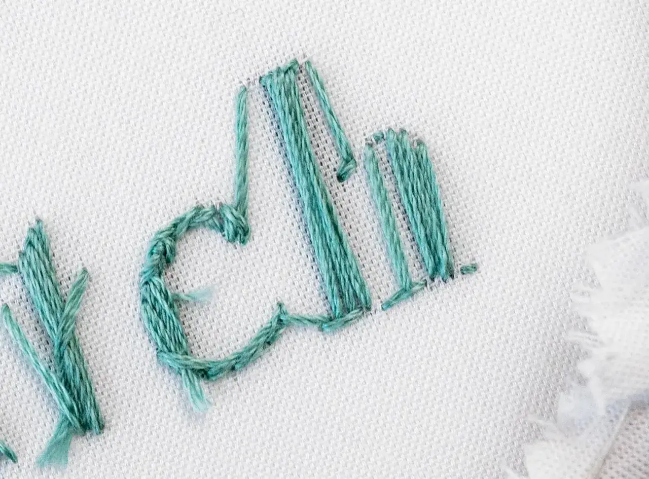 Detalle de letras bordadas con hilo turquesa sobre tela blanca. El proceso de bordar letras es delicado y detallado.