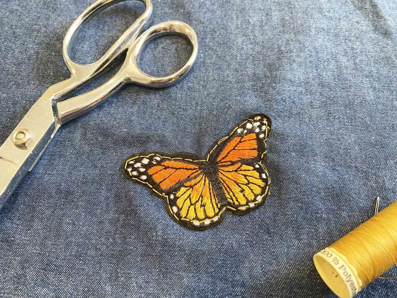 Un parche de mariposa bordado, tijeras y hilo amarillo sobre tela vaquera. Ideas para como hacer parches para ropa.