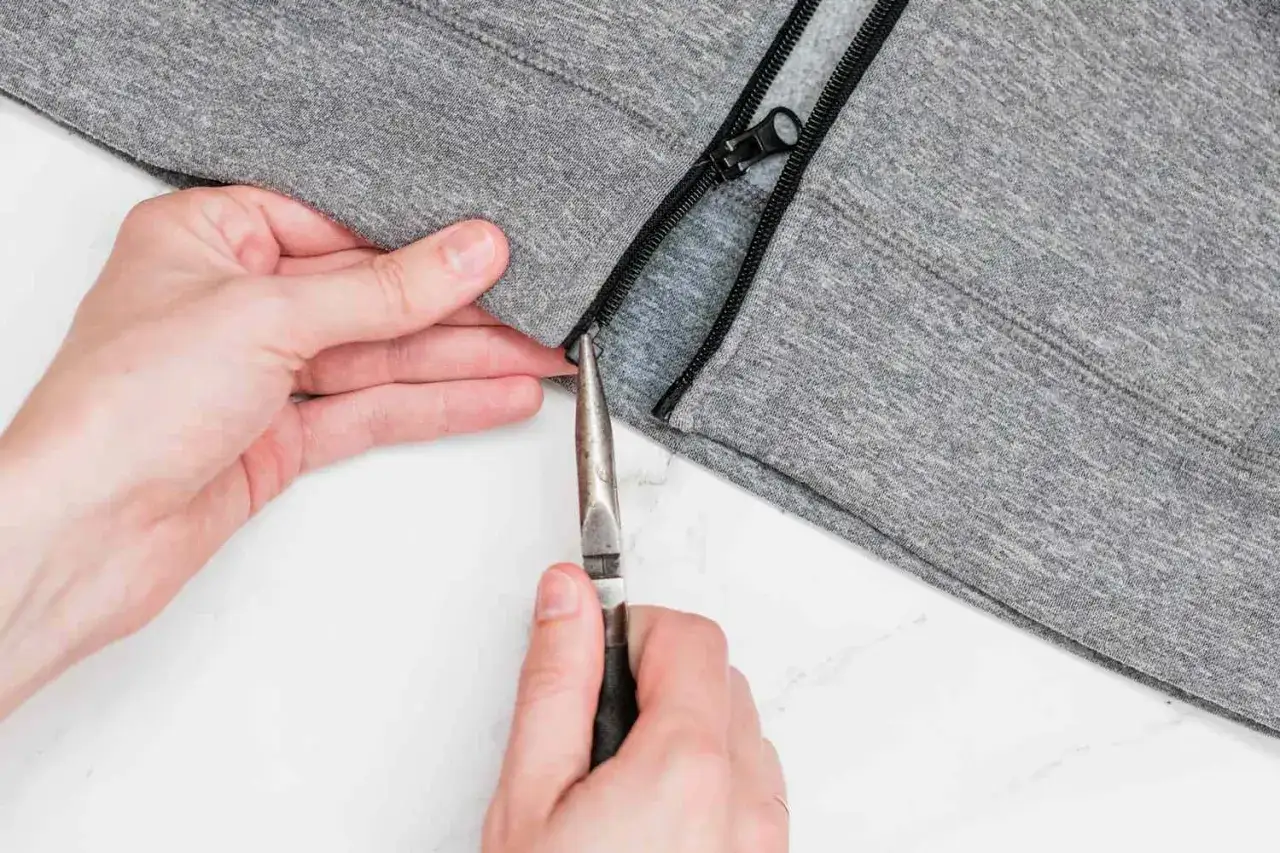 Manos intentan liberar una cremallera atascada con tela usando alicates en una sudadera gris.