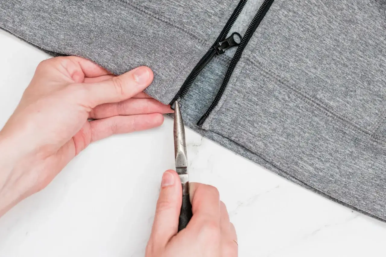 Manos arreglan una cremallera dura en una prenda gris con alicates.