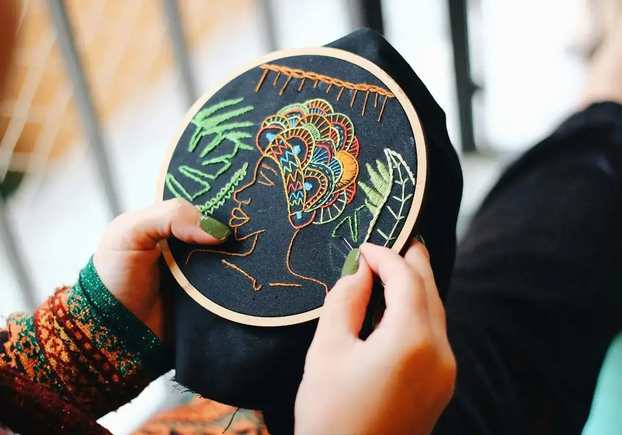 Manos creando arte con bordados modernos. Un rostro estilizado con un tocado vibrante sobre tela negra, rodeado de hojas verdes.