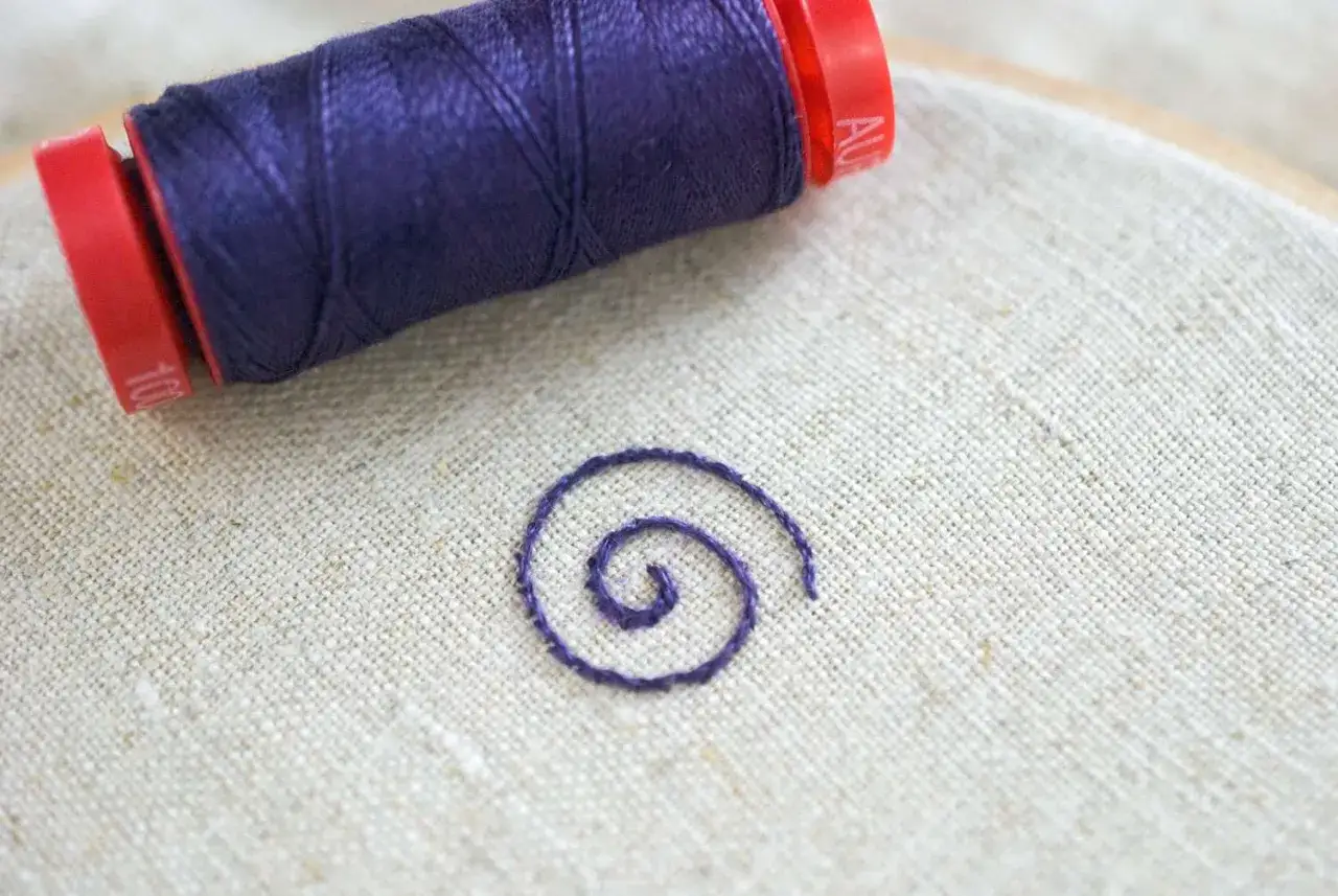 Un carretel de hilo de coser morado y un pequeño bordado en espiral sobre tela de lino.