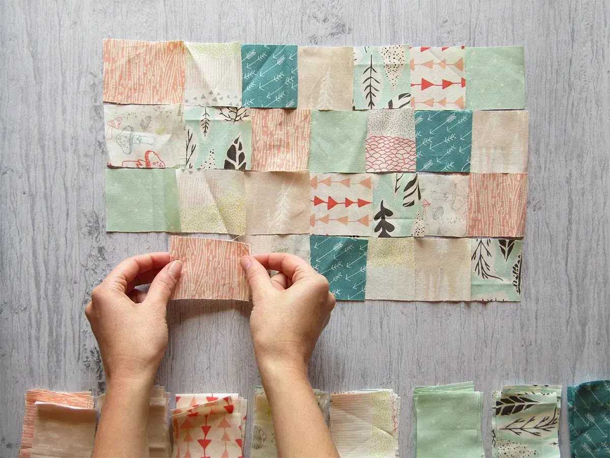 Manos uniendo retazos de tela con dise&ntilde;os de &aacute;rboles y flechas, creando un patr&oacute;n de patchwork.