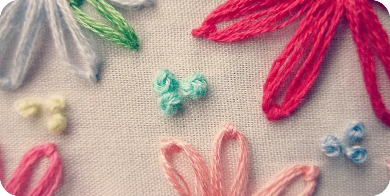 Detalle de bordado con flores de colores. Se aprecian p&eacute;talos en rosa, rojo, azul y blanco, y un grupo de nudos franceses bordados en turquesa.