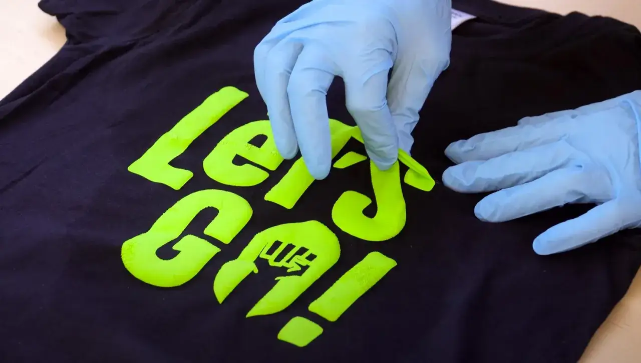 Manos enguantadas retirando una pegatina termoadhesiva verde ne&oacute;n de una camiseta negra. &iexcl;Un truco para quitar pegatinas termoadhesivas de la ropa!