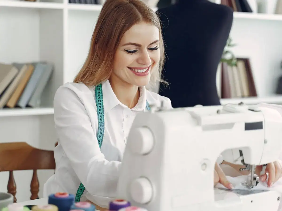 Mujer sonriente cosiendo ropa en máquina de coser, con cinta métrica al cuello.