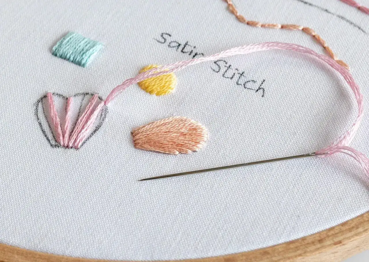 Bordado de muestra con hilo rosa y aguja. Se aprecian varios puntos, incluyendo el satin stitch en forma de pétalos y cuadrados.
