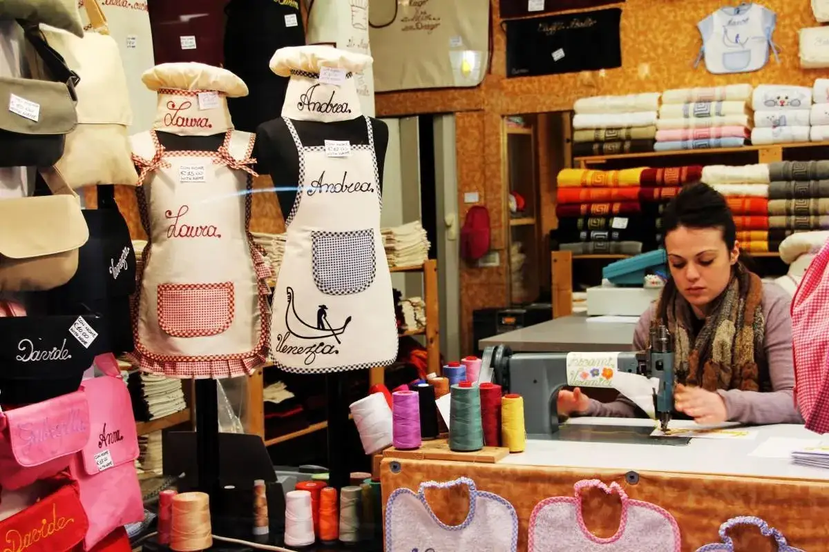 En esta mercer&iacute;a, delantales personalizados con nombres como Laura y Andrea, y bolsos bordados. Una mujer cose en una m&aacute;quina de coser.