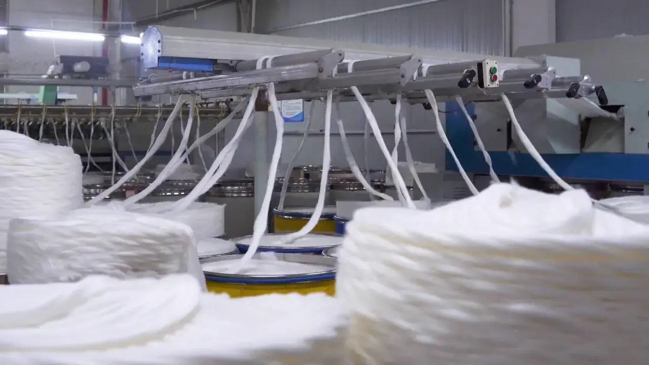 Máquinas textiles procesan grandes bobinas de algodón blanco, mostrando de donde sale el hilo que se transforma en tela.