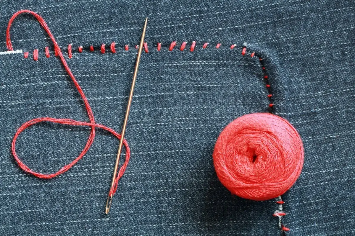 Hilo rojo, aguja y ovillo sobre tela vaquera, mostrando c&oacute;mo coser un agujero a mano con puntadas visibles.