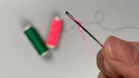Dedos sujetan una aguja para meter hilos, con carretes de hilo verde y rosa desenfocados al fondo.
