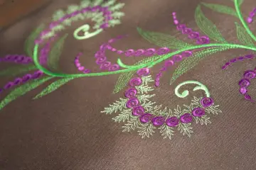 Detalles de bordado floral en tonos verdes y morados sobre tela marrón. Ideal para descargar letras para bordar a maquina y crear diseños únicos.