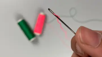 Dedos sujetan una aguja para meter hilos, con carretes de hilo verde y rosa desenfocados al fondo.