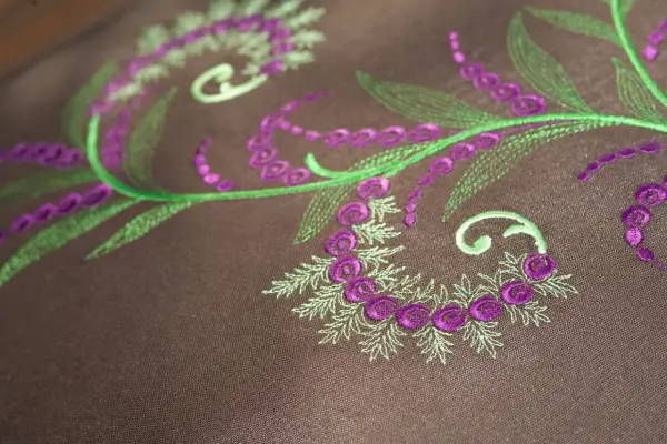 Detalles de bordado floral en tonos verdes y morados sobre tela marrón. Ideal para descargar letras para bordar a maquina y crear diseños únicos.