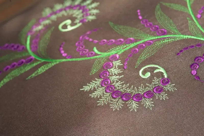 Detalles de bordado floral en tonos verdes y morados sobre tela marrón. Ideal para descargar letras para bordar a maquina y crear diseños únicos.