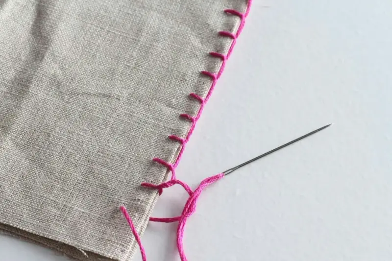 Primer plano de tela beige con puntadas a mano de hilo rosa brillante. Se aprecian varios tipos de puntadas a mano, incluyendo una puntada de festón.