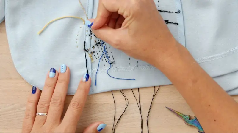 Manos con uñas decoradas bordando con hilo de coser sobre tela azul claro. Tijeras de colores y hilos negros a la vista.